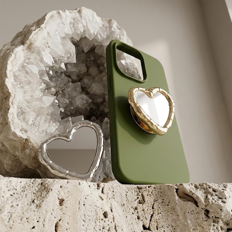May include: An olive green phone case with a gold-rimmed heart-shaped phone grip. A silver heart-shaped compact mirror is placed beside the phone case. The background features a geode and a textured surface.