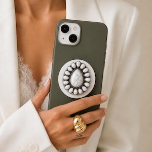 Pode incluir: Uma capa de smartphone verde-oliva com um suporte circular. O suporte tem uma pedra branca central em forma de gota com veios cinzentos, rodeada por uma borda de pedras brancas ovais mais pequenas numa moldura de metal escuro.