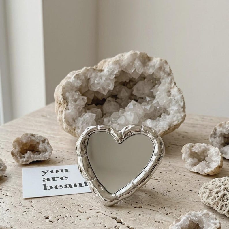 May include: A heart-shaped silver mirror with a decorative frame. The mirror is on a light-coloured surface, with a geode and small crystals in the background. A card with the words "you are beautiful" is also present.