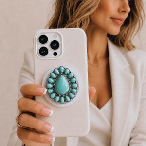 Puede incluir: Una funda de smartphone beige con un soporte circular para teléfono. El accesorio está decorado con una gran piedra turquesa central en forma de lágrima, rodeada por un anillo de piedras turquesas ovaladas más pequeñas engastadas en metal plateado.