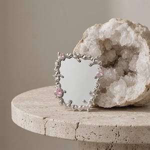 May include: A small, decorative silver mirror with an unusual, flowing frame. The frame is adorned with pink gemstones. The mirror is placed on a light-coloured, textured surface, with a geode in the background.