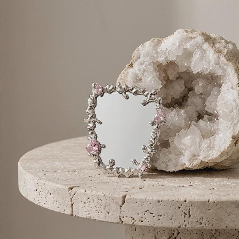 May include: A small, decorative silver mirror with an unusual, flowing frame. The frame is adorned with pink gemstones. The mirror is placed on a light-coloured, textured surface, with a geode in the background.