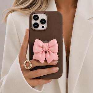 Op de afbeelding: Een donkerbruine smartphonehoes met een opvallende, glanzende lichtroze strik in het midden. De hoes heeft een uitsparing voor een camerasysteem met drie lenzen en een flitser, en biedt een gladde, matte afwerking voor de telefoon.