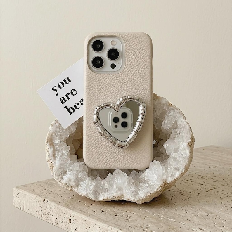 May include: A cream-coloured phone case featuring a heart-shaped mirror. The case has a textured finish and a silver frame. A small card with the words "you are be?" is placed near the phone, adding a decorative touch.