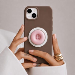 May include: A matte brown smartphone case featuring a circular white and light pink button-shaped grip attached to the center. The grip has a raised pink center with four small holes, resembling a classic sewing button, mounted on a white base.