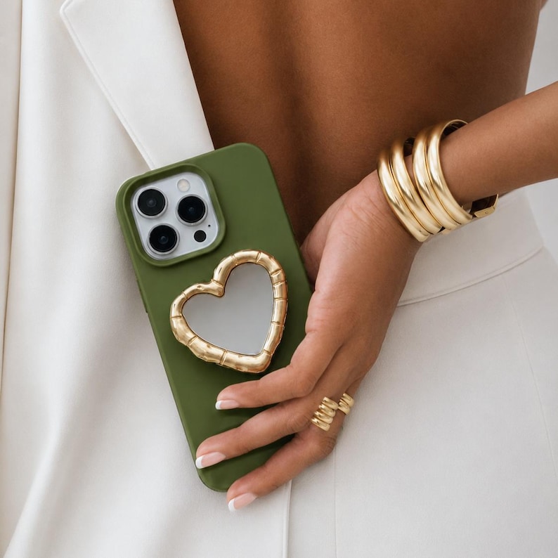 May include: Olive green phone case with a gold heart-shaped mirror. The case is held by a person wearing gold jewellery, including a thick bracelet and rings. The background is a white blazer and trousers.