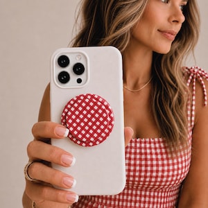 May include: A white smartphone case featuring a circular, collapsible phone grip with a red and white gingham checkered pattern. The phone case has a cutout for a triple-lens camera system on the back, providing a secure hold for the device.