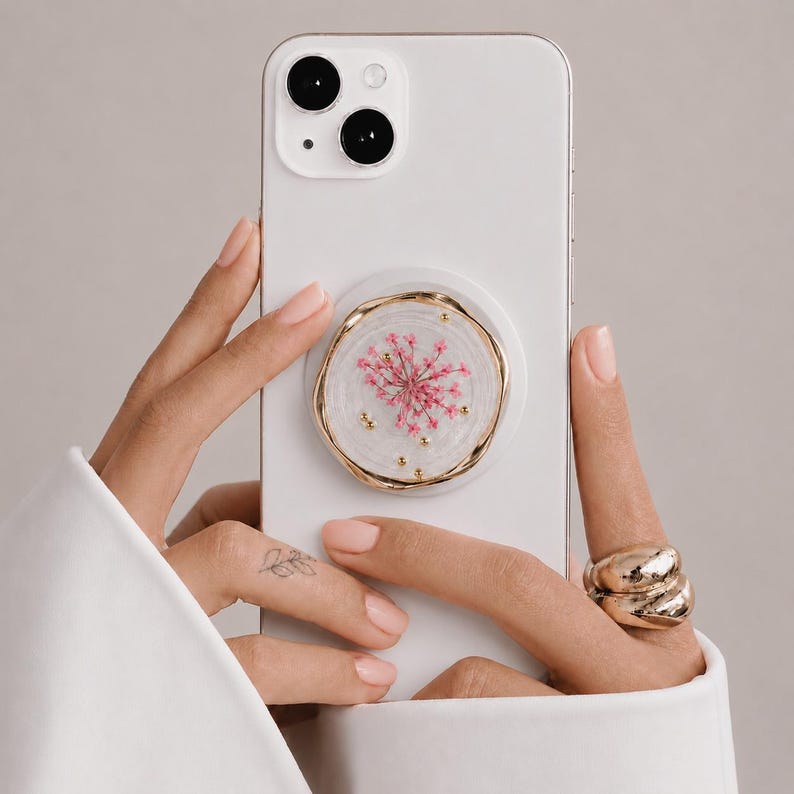 May include: A white smartphone featuring a circular phone grip attached to the back. The grip has a clear, resin-like finish containing a pressed pink flower and small gold-colored metallic beads, all encased within an irregular gold-toned rim.