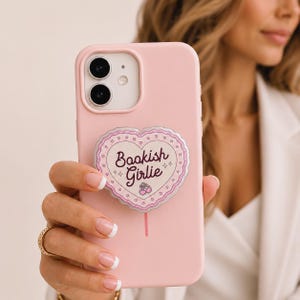 May include: A pink phone case with a heart-shaped pop socket that reads "Bookish Girlie." The pop socket has a silver border and a pink lollipop stick. The phone case is held in a hand with a gold ring.