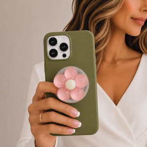 May include: An olive green smartphone case featuring a circular phone grip attachment on the back. The grip is designed as a five-petaled flower with translucent pink petals and a solid cream-colored center, positioned centrally on the case.