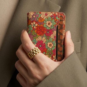 May include: A colourful floral wallet featuring red, pink, orange, and green flowers. The wallet has a brown interior and is held by a person wearing a gold ring. The background is a muted green.