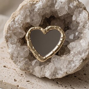 May include: A heart-shaped mirror with a gold-coloured bamboo-style frame. The mirror is set inside a geode with quartz crystals. The geode sits on a light-coloured, textured surface.