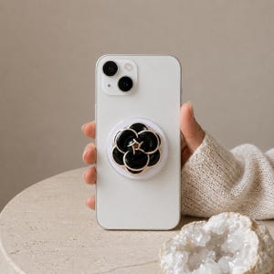 以下が含まれることがあります： 背面に円形のスマホグリップが付いた白いスマートフォン。グリップは光沢のある花びらと細い金色の金属製の縁取りを持つ黒い花のデザインで、スマートフォンの表面に立体的な層状の外観を作り出しています。