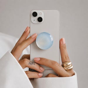 May include: A light gray smartphone featuring a circular, translucent, pale blue collapsible grip attached to the center of the back panel. The device has a dual-lens camera system in the upper left corner and a smooth, matte finish on the casing.