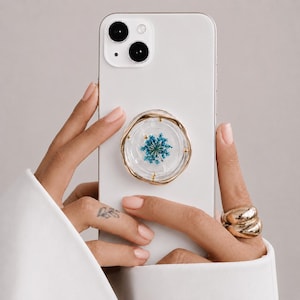 Pode incluir: Um smartphone branco com um suporte circular fixado na parte traseira. O suporte é feito de resina transparente com uma borda ondulada dourada e contém uma única flor azul prensada no centro, acentuada com pequenas partículas metálicas douradas.