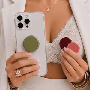 Op de afbeelding: Een doorzichtig smartphonehoesje met een olijfgroene, ronde, inklapbare grip aan de achterkant. Twee extra ronde grips in donkerbordeaux en oudroze worden apart in de andere hand gehouden voor een kleurvergelijking.