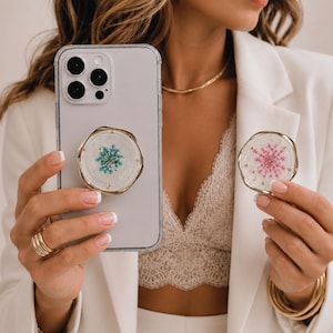 Peut inclure: Un étui de téléphone transparent avec un support de téléphone à bordure dorée, avec un fond blanc et un motif floral bleu. Un deuxième support de téléphone avec un motif floral rose est également visible. Les supports sont tenus par une personne.