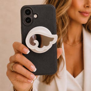 Könnte beinhalten: Ein schwarzes Smartphone mit einem weißen, flügelförmigen Handyhalter und einem kleinen Spiegel. Das Telefon hat eine Dual-Kamera. Der Halter ist an der Rückseite des Telefons befestigt.