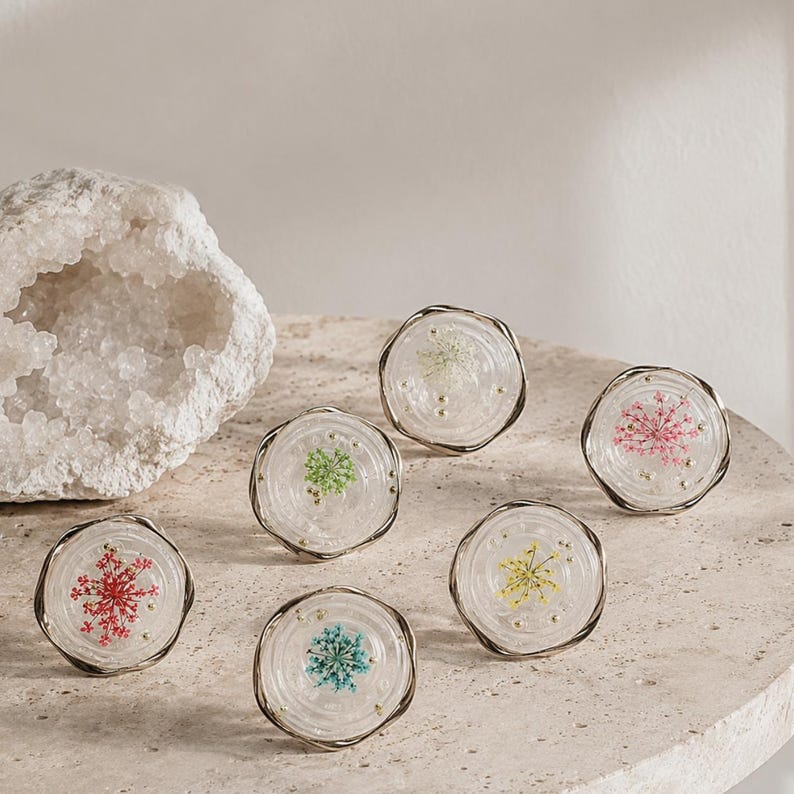 May include: Six circular cabinet knobs featuring clear resin centers with embedded dried flowers in red, green, white, pink, yellow, and blue, each encased in a thin metallic rim with small gold-toned accents throughout the clear material.
