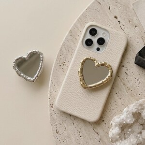 May include: A cream phone case with a gold heart-shaped mirror. A separate silver heart-shaped mirror is also visible. The mirrors have a textured border. The phone case is on a textured surface.