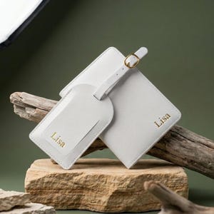 May include: A light gray luggage tag and passport holder set. The tag and holder are made of a smooth material and feature the name "Lisa" in gold lettering. The tag has a white strap with a gold buckle. The set is displayed on a stone surface.
