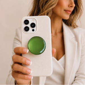 Peut inclure: Un étui de téléphone crème avec une poignée circulaire verte. L'étui du téléphone a un appareil photo à trois objectifs et est tenu par une personne portant un blazer blanc et des bijoux en or.