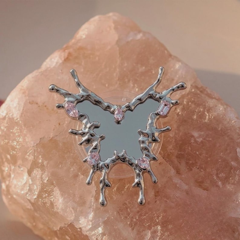 May include: A silver butterfly-shaped accessory with a mirrored centre and pink gemstone accents. The butterfly has an irregular, melted metal design. The accessory is set against a light pink, textured background.