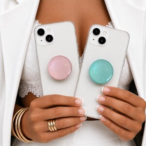 May include: Two clear smartphone cases, each featuring a circular collapsible grip on the back. One grip is a solid pastel pink, and the other is a solid pastel teal. Both cases have cutouts for dual rear camera lenses and a flash.