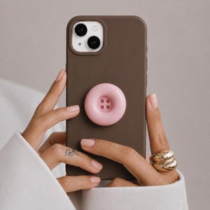 May include: A dark brown smartphone case featuring a circular, light pink, four-hole button-shaped grip attached to the center of the back. The case has a raised camera module cutout at the top left corner, revealing two camera lenses.