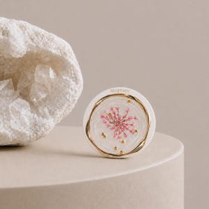 May include: A circular white magnetic object featuring a clear resin center embedded with a pink dried flower and small gold-colored metallic beads. The word Magnetic is printed in small letters at the top edge of the white border.