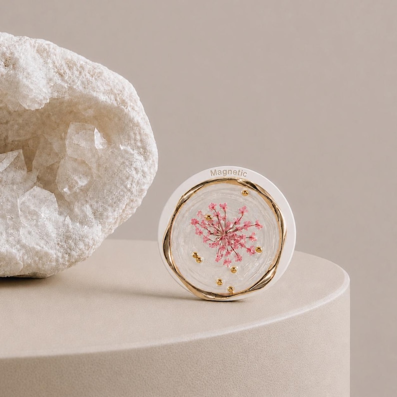 May include: A circular white magnetic object featuring a clear resin center embedded with a pink dried flower and small gold-colored metallic beads. The word Magnetic is printed in small letters at the top edge of the white border.