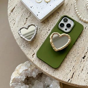 May include: A silver heart-shaped mirror and a gold heart-shaped phone grip are displayed on a textured surface. An olive green phone case with a gold heart-shaped mirror grip is also shown. A white tray with gold rings and a pearl necklace are in the background.