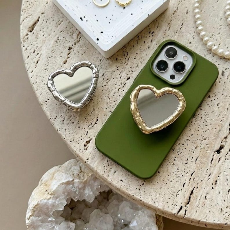 May include: A silver heart-shaped mirror and a gold heart-shaped phone grip are displayed on a textured surface. An olive green phone case with a gold heart-shaped mirror grip is also shown. A white tray with gold rings and a pearl necklace are in the background.