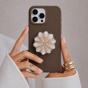 Op de afbeelding: Een mat donkerbruin smartphonehoesje met een camera met drie lenzen en een ronde, doorschijnende wit-gouden bloemvormige gripbevestiging in het midden, met een gelaagd bloembladontwerp en een klein accent in het midden.