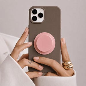Op de afbeelding: Een mat donkerbruin smartphonehoesje met een ronde, lichtroze inklapbare grip in het midden van de achterkant. Het hoesje heeft een verhoogde uitsparing voor de camera met drie zichtbare ronde lenzen. Het ontwerp is eenvoudig en functioneel.