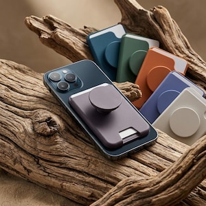 Puede incluir: Un teléfono inteligente azul oscuro con un agarre para teléfono y un tarjetero morados. También se ven varios agarres para teléfono y tarjeteros de colores en azul, verde, naranja, azul claro y blanco, todos sobre un trozo de madera flotante.