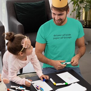 May include: A teal t-shirt with the text "driver of kids payer of things" is worn by a person wearing a paper crown. A child is drawing with markers on paper. The scene is set on a black table.