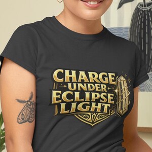 May include: A dark gray short-sleeved t-shirt featuring a gold graphic print on the chest. The text reads "CHARGE UNDER ECLIPSE LIGHT" in a bold, stylized font, accompanied by geometric shapes, star accents, and a vertical rectangular emblem.
