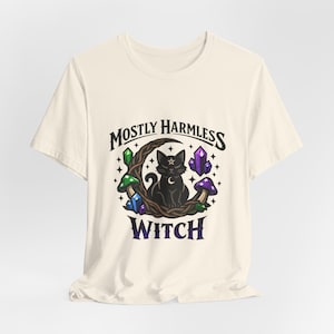 May include: Cream-colored t-shirt featuring a black cat with a star on its head, sitting on a crescent moon. The design includes crystals, mushrooms, and the text "MOSTLY HARMLESS WITCH" in a gothic font. The shirt is made of a soft material.