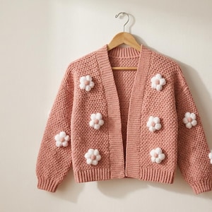 May include: A dusty rose open-front cardigan featuring a textured knit pattern and white pom-pom flower appliqués on the front and sleeves. The garment has long sleeves with ribbed cuffs, a ribbed hem, and a ribbed collar, all hanging on a wooden hanger.