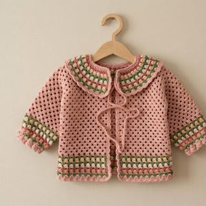 May include: A light pink crochet baby cardigan with a scalloped collar and cuffs. The collar and cuffs feature a striped pattern in shades of green, pink, and cream. The cardigan has a tie closure and is displayed on a wooden hanger.