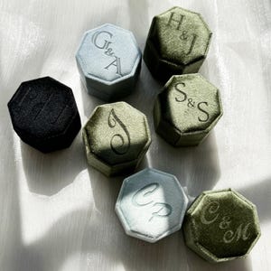 May include: Seven octagonal velvet ring boxes in black, light blue, and olive green. Each box features a unique monogram or set of initials embossed on the lid, including G&A, H&J, S&S, J, CP, and C&M, with one black box showing faint lettering.