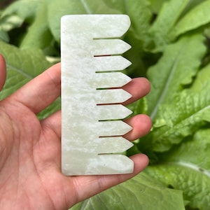 May include: A light green jade comb with a rectangular shape and pointed teeth. The comb is held in a hand against a background of green leaves. The comb is a beauty tool.
