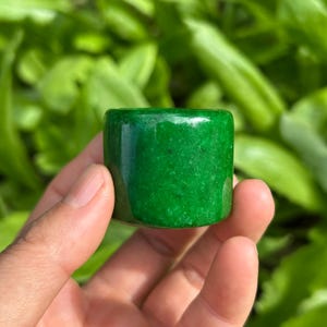 May include: A cylindrical ring band crafted from a solid, polished green stone with a smooth, glossy finish. The material displays natural variations in color intensity and texture throughout its surface, showcasing a vibrant, deep emerald hue.