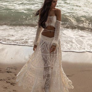 May include: A cream crochet two-piece outfit on a beach. The outfit features a strapless top with a lace-up design, long sleeves, and a flowing skirt. The intricate crochet pattern is visible throughout the ensemble.