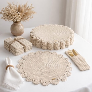 Puede incluir: Un conjunto de manteles individuales y posavasos de ganchillo de color crema. Los manteles individuales tienen un diseño de borde festoneado. Los posavasos están apilados y atados con hilo. También hay un portacubiertos a juego y una servilleta con un anillo de madera.