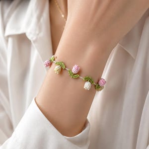 Può includere: Un braccialetto delicato caratterizzato da piccoli fiori di tulipano all'uncinetto in tonalità rosa tenue, crema e bianco, intervallati da foglie verdi all'uncinetto lungo un sottile cinturino flessibile che avvolge il polso.