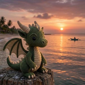 以下が含まれることがあります： 大きな黒い目をした、手作りの緑とベージュのかぎ針編みのドラゴンが木の柱に座っています。ドラゴンは詳細な翼と角を持っています。背景には、海に沈む夕日と小さなボートがあるビーチのシーンがあります。