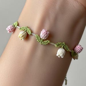 Può includere: Un delicato braccialetto all'uncinetto con un design floreale. Il braccialetto presenta fiori all'uncinetto rosa, bianchi e gialli con foglie verdi. Il braccialetto è realizzato con una sottile catena color argento.