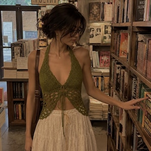May include: A person wearing a green crochet halter top and a cream-colored skirt with a floral pattern. The person is in a bookstore, browsing books on a shelf. The word "POETRY" is visible above.